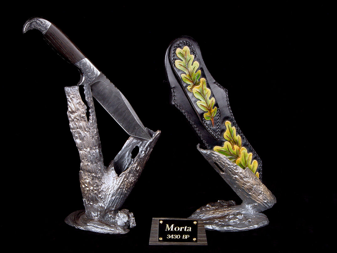 "Morta" reverse side view in twist damascus pattern welded steel blade, hand-engraved 304 stainless steel bolsters, Bog Oak mineralized hardwood handle, sheath of hand-carved leather shoulder, hand-dyed, stand of hand-cast bronze